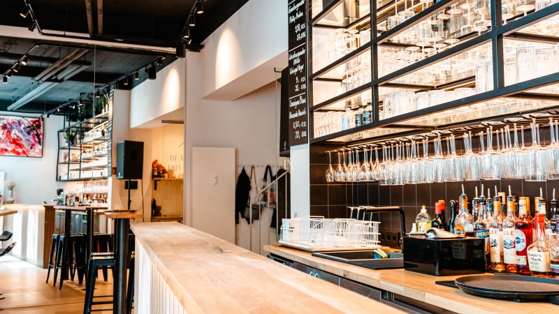 Moderne Bar mit hölzernem Tresen, Regalen voller Gläser und Flaschen. Helle Beleuchtung und stilvolle Einrichtung schaffen eine einladende Atmosphäre., © SMG_Sarah Schmid
