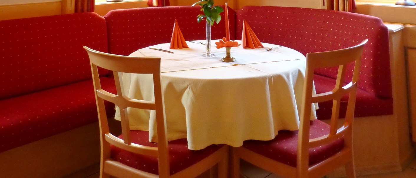 Cozy restaurant corner with round table, white tablecloth, red chairs and curtains. There is a vase of flowers on the table., © Hotel & Restaurant Sonne