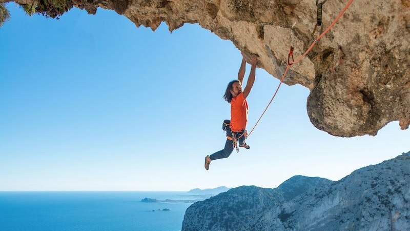 Ein Kletterer in orangefarbenem Shirt hängt an einer steilen Felswand über dem Meer, gesichert mit einem Seil. Im Hintergrund ist das Meer zu sehen., © Theaterhaus Stuttgart e.V. Ein Kletterer in orangefarbenem Shirt hängt an einer steilen Felswand über dem Meer, gesichert mit einem Seil. Im Hintergrund ist das Meer zu sehen., © Theaterhaus Stuttgart e.V.
