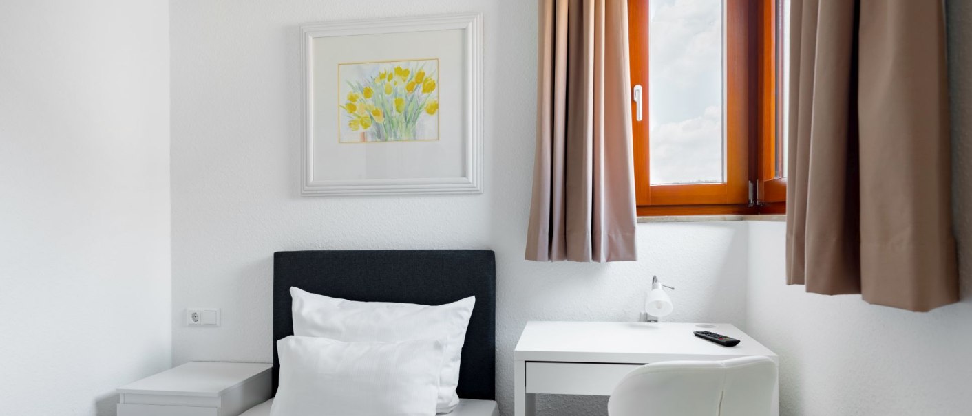 Bright single room with a bed, a white desk and a window with brown curtains. A picture of yellow flowers hangs on the wall., © Hotel Olgaeck Bright single room with a bed, a white desk and a window with brown curtains. A picture of yellow flowers hangs on the wall., © Hotel Olgaeck