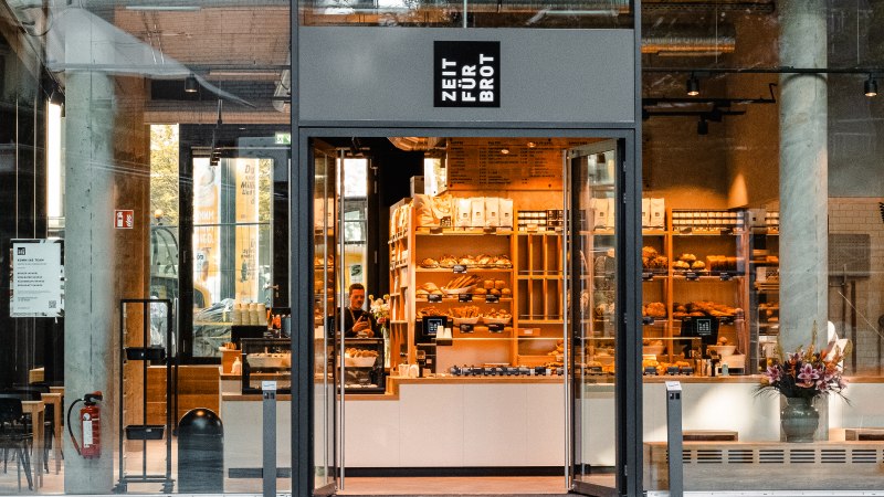 Entrance to a bakery with a glass front. Shelves with baked goods can be seen inside. 'Time for bread' is written above the door., &copy; Zeit f&uuml;r Brot Stuttgart