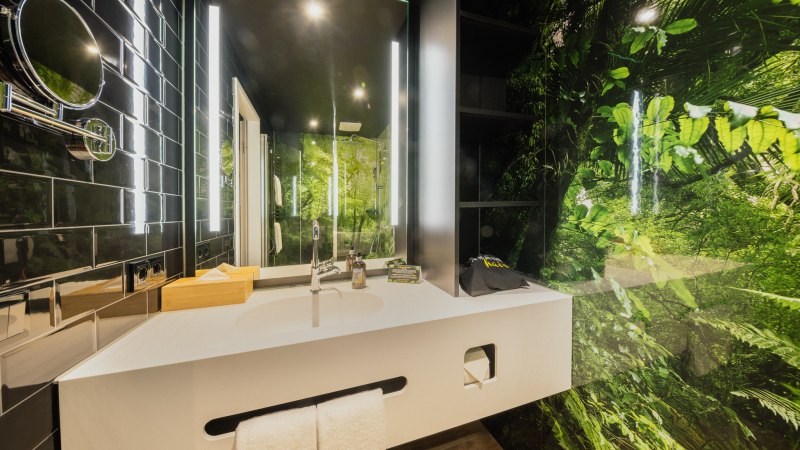 Modernes Badezimmer mit weißem Waschbecken, Spiegel und Dschungelmotiv an der Wand. Schwarze Fliesen und helle Beleuchtung ergänzen das Design., © Rioca Stuttgart Posto 4 GmbH Modernes Badezimmer mit weißem Waschbecken, Spiegel und Dschungelmotiv an der Wand. Schwarze Fliesen und helle Beleuchtung ergänzen das Design., © Rioca Stuttgart Posto 4 GmbH