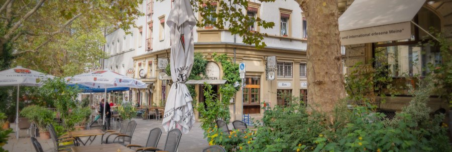 A bistro on Wilhelmsplatz with empty tables and chairs outside, surrounded by green plants and trees., &copy; Martina Denker