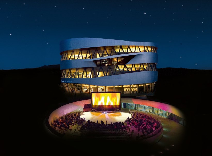 A modern, illuminated building at night. In the foreground, an open-air cinema with spectators and a large screen., © Mercedes-Benz Heritage GmbH A modern, illuminated building at night. In the foreground, an open-air cinema with spectators and a large screen., © Mercedes-Benz Heritage GmbH