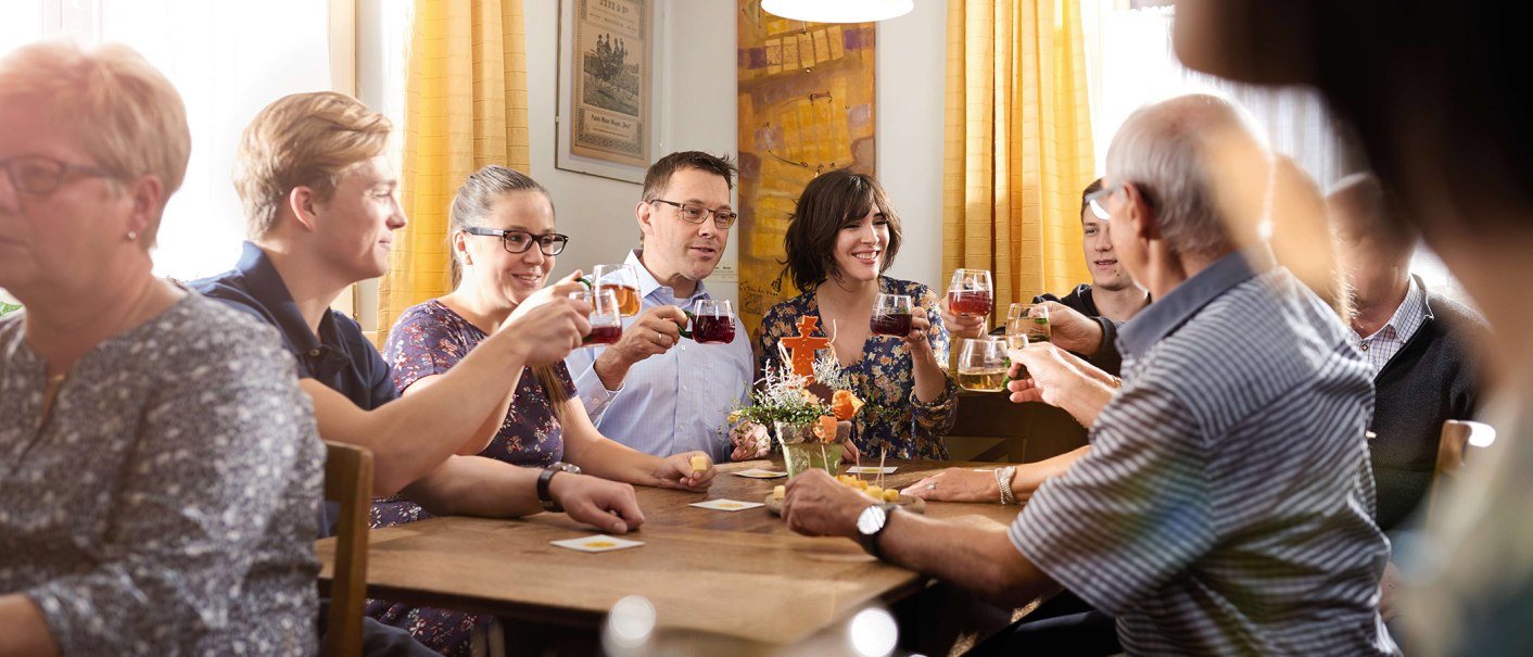 Eine Gruppe von Menschen sitzt an einem Tisch und st&ouml;&szlig;t mit Getr&auml;nken an. Sie wirken fr&ouml;hlich und entspannt. Im Hintergrund sind gelbe Vorh&auml;nge., &copy; Stuttgart-Marketing GmbH / Winkler
