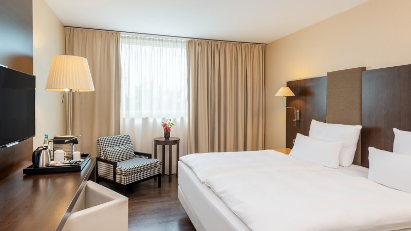 Modernes Doppelzimmer mit gro&szlig;em Bett, Schreibtisch, Sessel und Fenster mit beigen Vorh&auml;ngen. Ein Fernseher und eine Stehlampe sind ebenfalls vorhanden., &copy; NH Hotel Stuttgart Airport