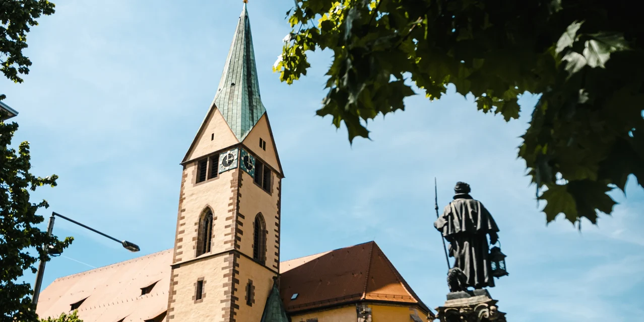 Leonardskirche in Stuttgart mit Turm, Uhr und Statue des Nachtwächterbrunnens im Vordergrund, umrahmt von grünen Blättern unter blauem Himmel., © SMG, Sarah Schmid Leonardskirche in Stuttgart mit Turm, Uhr und Statue des Nachtwächterbrunnens im Vordergrund, umrahmt von grünen Blättern unter blauem Himmel., © SMG, Sarah Schmid