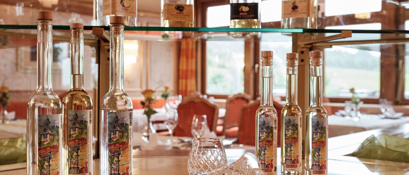 Flaschen mit Etiketten und Gläser auf einem Tisch in einem eleganten Restaurant. Im Hintergrund sind gedeckte Tische und große Fenster zu sehen., © Badhotel Restaurant Stauferland