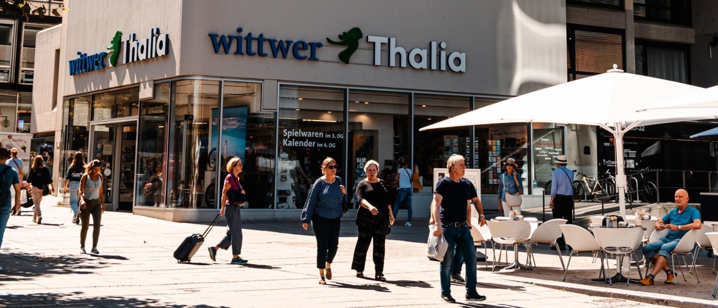 Menschen flanieren vor der Buchhandlung Wittwer-Thalia. Einige sitzen unter Sonnenschirmen in einem Café. Es ist ein sonniger Tag., © Stuttgart Marketing GmbH, Sarah Schmid
