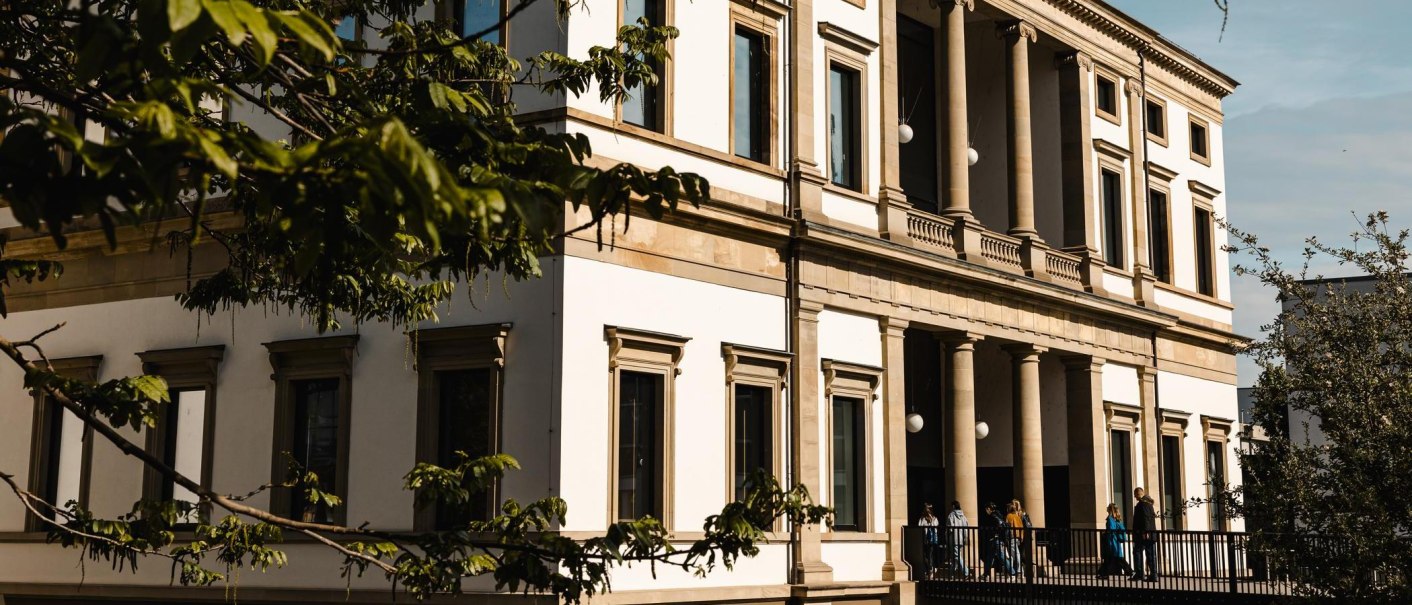 A historic building with columns and large windows, surrounded by trees. People walk on a path along the façade., © SMG Stuttgart Marketing GmbH - Sarah Schmid A historic building with columns and large windows, surrounded by trees. People walk on a path along the façade., © SMG Stuttgart Marketing GmbH - Sarah Schmid