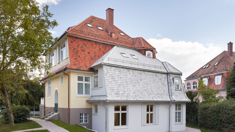 Zweistöckiges Haus mit rotem Ziegeldach, gelber Fassade und weißen Fenstern, umgeben von Bäumen und Rasen., © Philip Kottlorz Zweistöckiges Haus mit rotem Ziegeldach, gelber Fassade und weißen Fenstern, umgeben von Bäumen und Rasen., © Philip Kottlorz