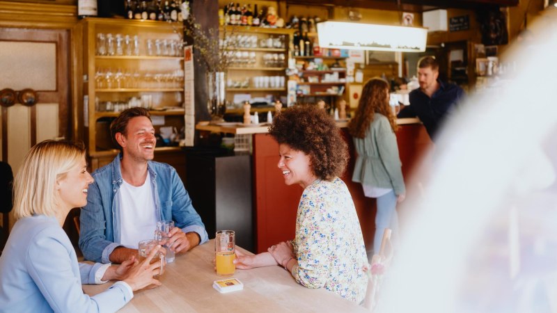 Three people sit laughing at a table in a cozy bar. Glasses and bottles can be seen in the background., © © Stuttgart-Marketing GmbH, Alwin Maigler Three people sit laughing at a table in a cozy bar. Glasses and bottles can be seen in the background., © © Stuttgart-Marketing GmbH, Alwin Maigler
