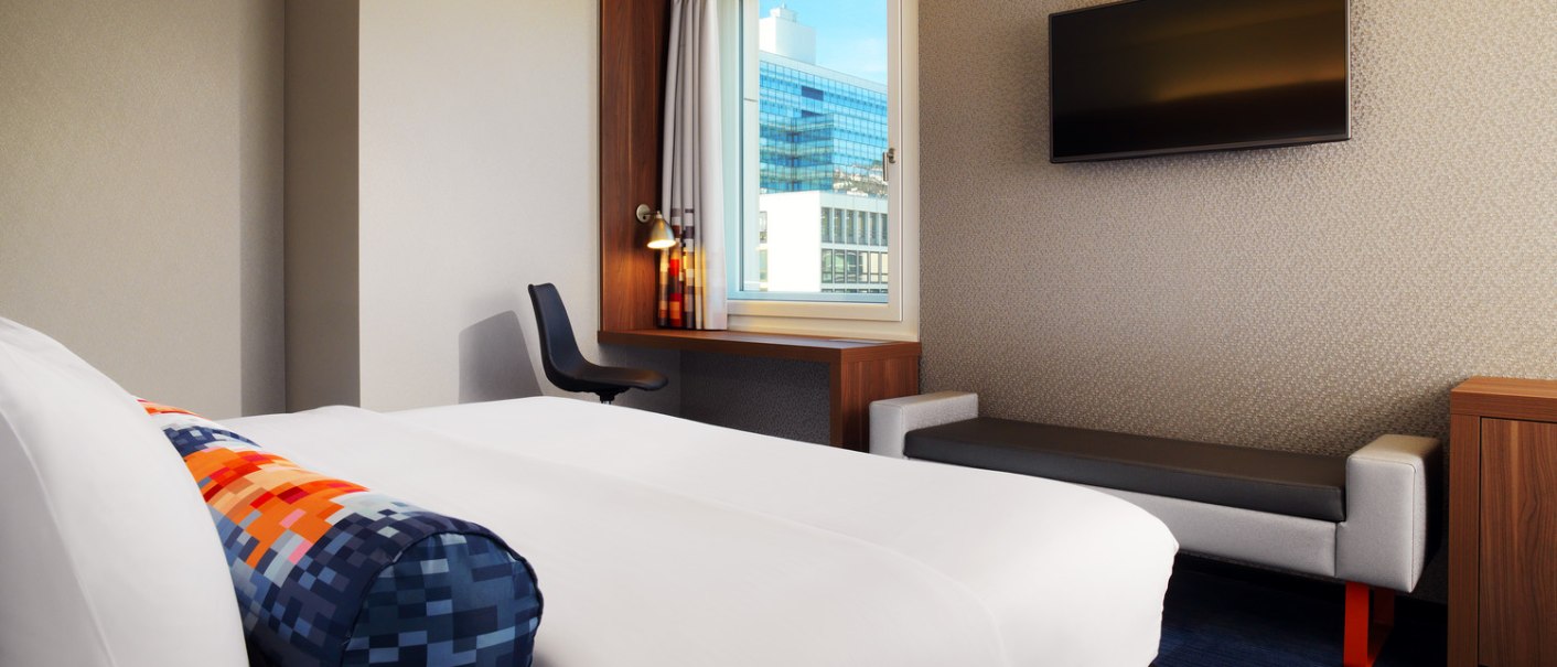 Ein modernes Hotelzimmer mit einem großen Bett, einem Schreibtisch am Fenster und einem Flachbildfernseher an der Wand., © Aloft Stuttgart Ein modernes Hotelzimmer mit einem großen Bett, einem Schreibtisch am Fenster und einem Flachbildfernseher an der Wand., © Aloft Stuttgart