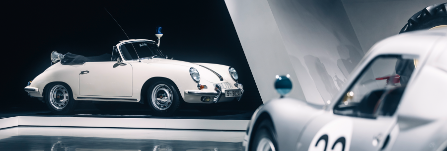 Two classic cars in an exhibition. A white convertible with police lights and a silver car with the number 33 in the foreground., © Porsche AG Two classic cars in an exhibition. A white convertible with police lights and a silver car with the number 33 in the foreground., © Porsche AG