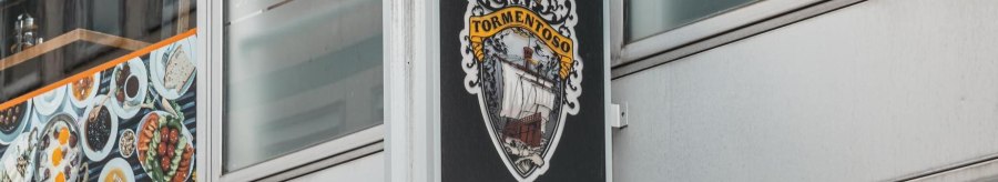A sign with the Cape Tormentoso logo hangs on a modern building. It shows a sailing ship and decorative elements., © Stuttgart Marketing GmbH, Sarah Schmid