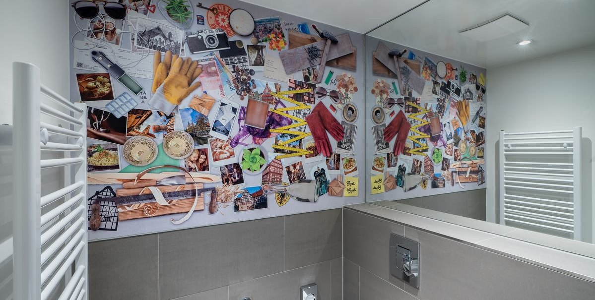 Badezimmer mit bunter Collage aus Fotos und Gegenständen an der Wand, reflektiert im Spiegel. Ein Handtuchhalter ist an der Seite sichtbar., © Novum Hospitality