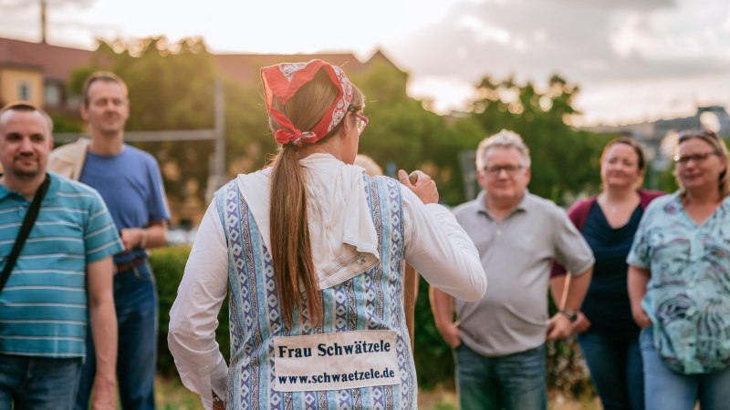 Eine Frau mit Kopftuch und traditioneller Kleidung spricht zu einer Gruppe von Menschen. Auf ihrem R&uuml;cken steht "Frau Schw&auml;tzele". Die Szene ist im Freien., &copy; &copy; Stuttgart-Marketing GmbH, Thomas Niederm&uuml;ller