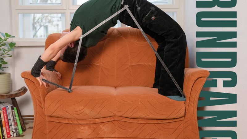 Eine Person macht eine Br&uuml;cke auf einem orangefarbenen Sofa, unterst&uuml;tzt von einem Metallgestell. Im Hintergrund sind Fenster und Pflanzen zu sehen., &copy; Merz Akademie