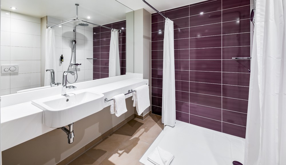 Modernes barrierefreies Badezimmer mit ebenerdiger Dusche, weißen und lila Fliesen, Waschbecken und Handtüchern., © Premier Inn Modernes barrierefreies Badezimmer mit ebenerdiger Dusche, weißen und lila Fliesen, Waschbecken und Handtüchern., © Premier Inn