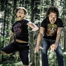 Zwei M&auml;nner springen energiegeladen in einem Wald. Sie tragen T-Shirts und Jeans und wirken fr&ouml;hlich und abenteuerlustig., &copy; links im Bild