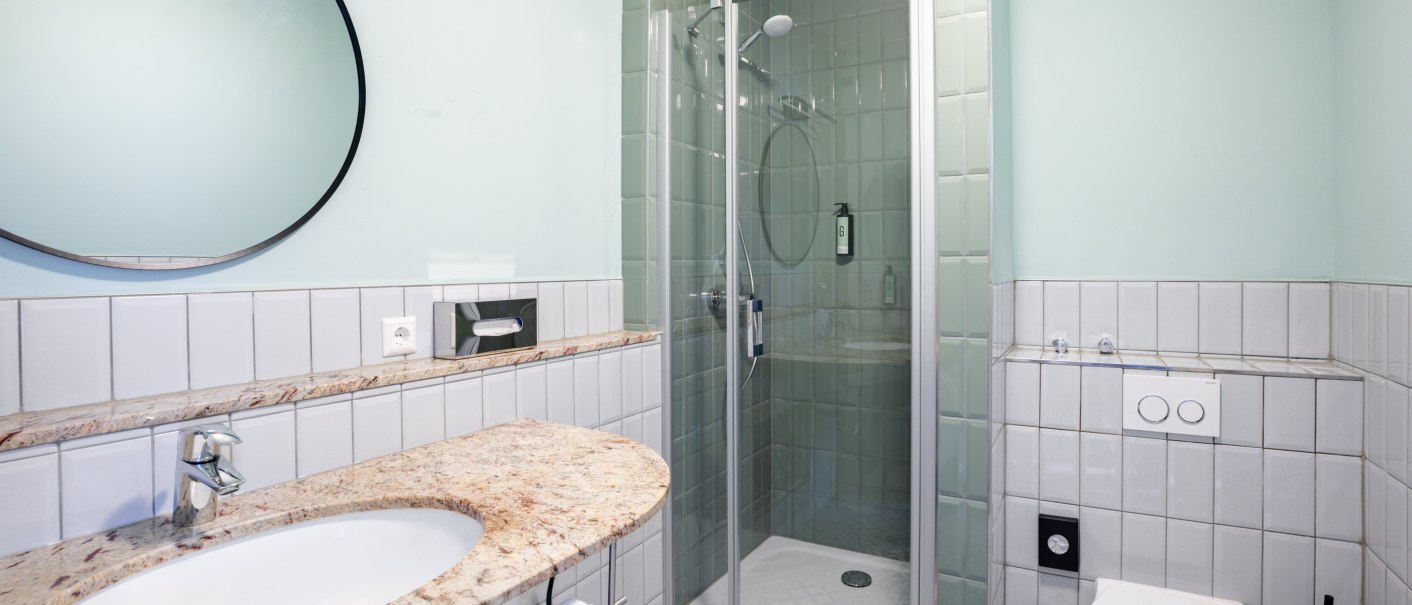 Ein modernes Badezimmer mit einer Glasdusche, einem runden Spiegel &uuml;ber einem Waschbecken und einer Toilette. Die W&auml;nde sind in Pastellfarben gehalten., &copy; Novum  Hospitality