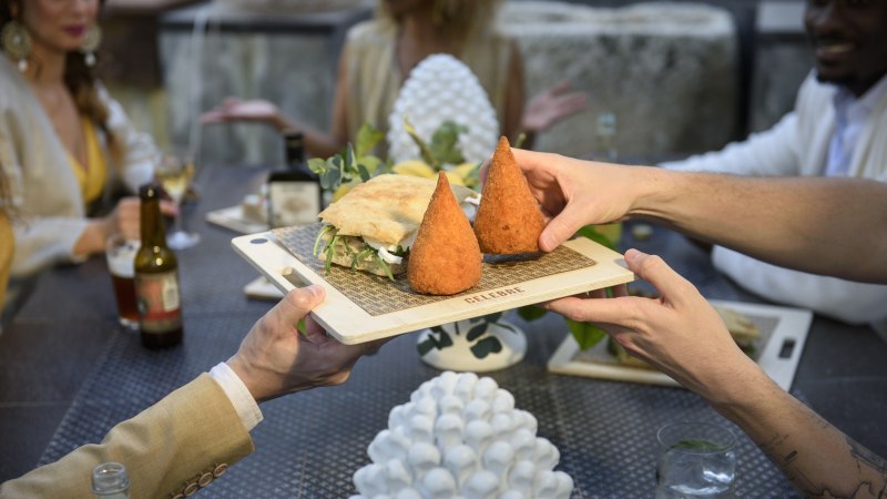Zwei Personen reichen ein Holzbrett mit Arancini und einem Sandwich an einem Tisch mit mehreren Gästen., © CELEBRE