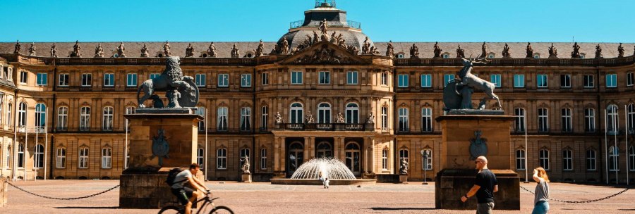 Das Neue Schloss Stuttgart bei sonnigem Wetter. Menschen gehen und ein Fahrradfahrer fährt im Vordergrund. Zwei Statuen flankieren den Eingang., © Stuttgart-Marketing GmbH, Sarah Schmid Das Neue Schloss Stuttgart bei sonnigem Wetter. Menschen gehen und ein Fahrradfahrer fährt im Vordergrund. Zwei Statuen flankieren den Eingang., © Stuttgart-Marketing GmbH, Sarah Schmid