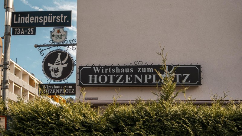 Sign for the Wirtshaus zum Hotzenplotz on Lindenspürstraße, surrounded by green bushes and a blue sky in the background., © SMG Stuttgart Marketing GmbH - Sarah Schmid