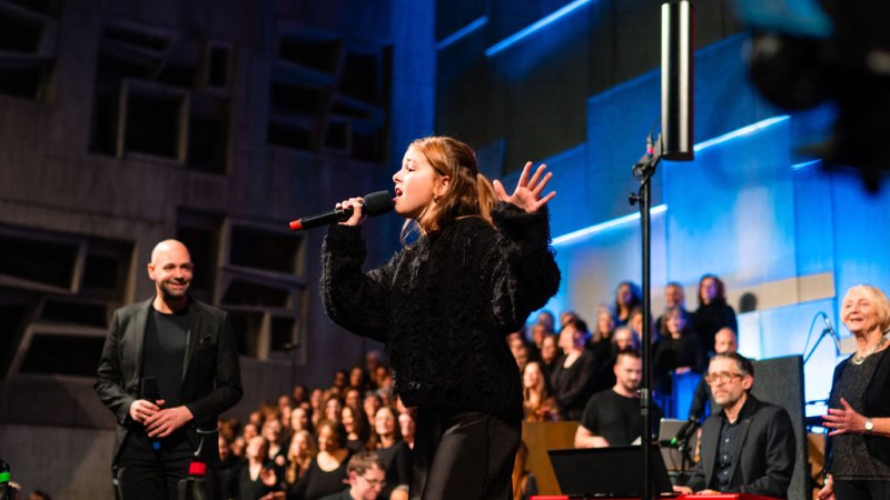 Ein Mädchen singt leidenschaftlich auf einer Bühne, begleitet von einem Chor und Musikern. Die Bühne ist in Blau- und Grautönen beleuchtet., © Ludmilla Parsyak Ein Mädchen singt leidenschaftlich auf einer Bühne, begleitet von einem Chor und Musikern. Die Bühne ist in Blau- und Grautönen beleuchtet., © Ludmilla Parsyak
