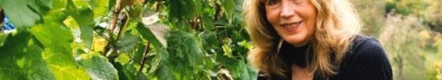 A woman with blonde hair harvests grapes in a green vineyard. She wears gloves and smiles at the camera., &copy; Elsbeth Koch