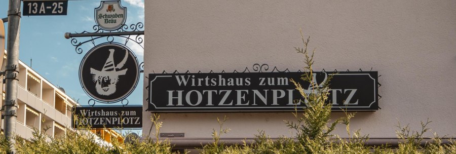 Schild des Wirtshauses zum Hotzenplotz an der Lindenspürstraße, umgeben von grünen Büschen und einem blauen Himmel im Hintergrund., © SMG. Sarah Schmid