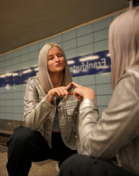 Frau mit silberner Jacke posiert vor einem Spiegel in einer U-Bahn-Station. Sie macht eine Handgeste und lächelt leicht., © Theaterhaus Stuttgart e.V. Frau mit silberner Jacke posiert vor einem Spiegel in einer U-Bahn-Station. Sie macht eine Handgeste und lächelt leicht., © Theaterhaus Stuttgart e.V.