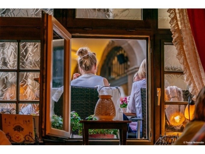 Blick durch ein offenes Fenster auf zwei Personen, die in einem gemütlichen Restaurant sitzen. Draußen sind Pflanzen und eine warme Beleuchtung zu sehen., © Stuttgart Marketing GmbH Blick durch ein offenes Fenster auf zwei Personen, die in einem gemütlichen Restaurant sitzen. Draußen sind Pflanzen und eine warme Beleuchtung zu sehen., © Stuttgart Marketing GmbH