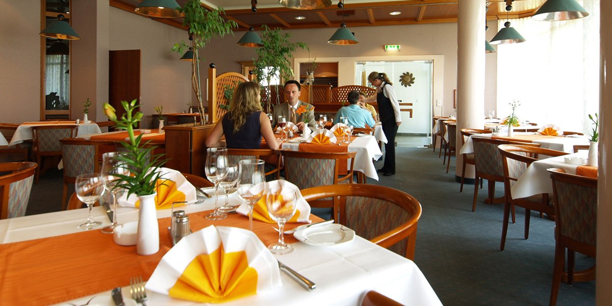 Ein elegantes Restaurant mit weiß gedeckten Tischen, orangefarbenen Servietten und Gästen, die speisen. Eine Kellnerin bedient einen Tisch., © © H-Hotels.com Ein elegantes Restaurant mit weiß gedeckten Tischen, orangefarbenen Servietten und Gästen, die speisen. Eine Kellnerin bedient einen Tisch., © © H-Hotels.com