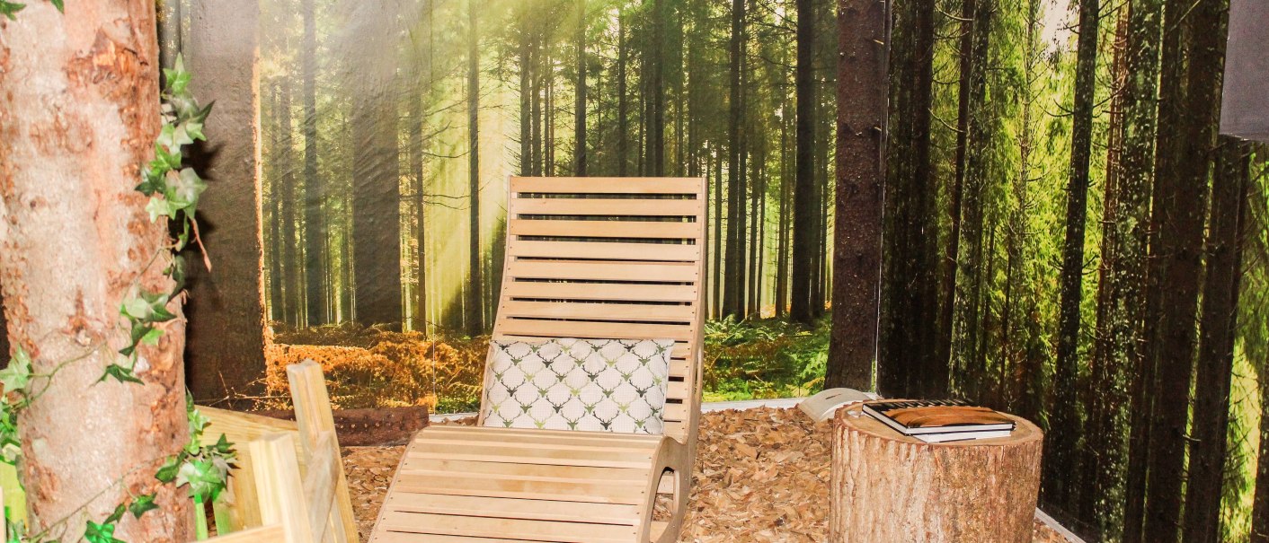 Wooden lounger in a room with forest wallpaper, sunbeams and wood shavings on the floor. A tree stump serves as a table., &copy; TOMAS