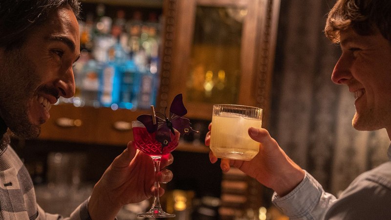 Zwei M&auml;nner sto&szlig;en in einer Bar mit Cocktails an, einer in einem karierten Sakko, der andere in einem hellen Hemd. Im Hintergrund sind Flaschen zu sehen., &copy; SMG, Martina Denker