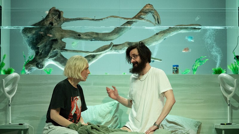 Zwei Personen sitzen auf einem Bett vor einem großen Aquarium und unterhalten sich. Im Hintergrund sind Fische und Pflanzen zu sehen., © Württembergische Staatstheater Stuttgart Zwei Personen sitzen auf einem Bett vor einem großen Aquarium und unterhalten sich. Im Hintergrund sind Fische und Pflanzen zu sehen., © Württembergische Staatstheater Stuttgart
