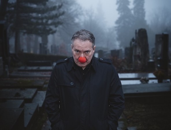 Eine Person mit roter Clownsnase steht auf einem nebligen Friedhof. Sie tr&auml;gt einen dunklen Mantel und hat die H&auml;nde in den Taschen., &copy; Renitenztheater Stuttgart e.V.