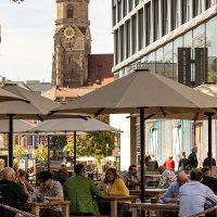 Menschen genie&szlig;en das sonnige Wetter im Dorotheenquartier in Stuttgart. Sie sitzen unter Sonnenschirmen, w&auml;hrend im Hintergrund eine Kirche zu sehen ist., &copy; Stuttgart-Marketing GmbH, Sarah Schmid