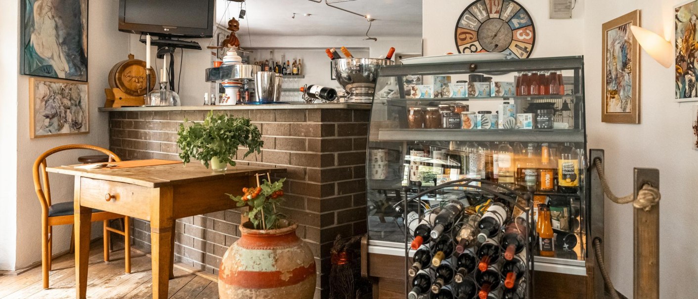 Gemütliches Café mit Holztisch, Weinregal, dekorativen Bildern und Pflanzen. Eine Theke mit Getränken und ein Fernseher sind ebenfalls zu sehen., © SMG, Sarah Schmid Gemütliches Café mit Holztisch, Weinregal, dekorativen Bildern und Pflanzen. Eine Theke mit Getränken und ein Fernseher sind ebenfalls zu sehen., © SMG, Sarah Schmid