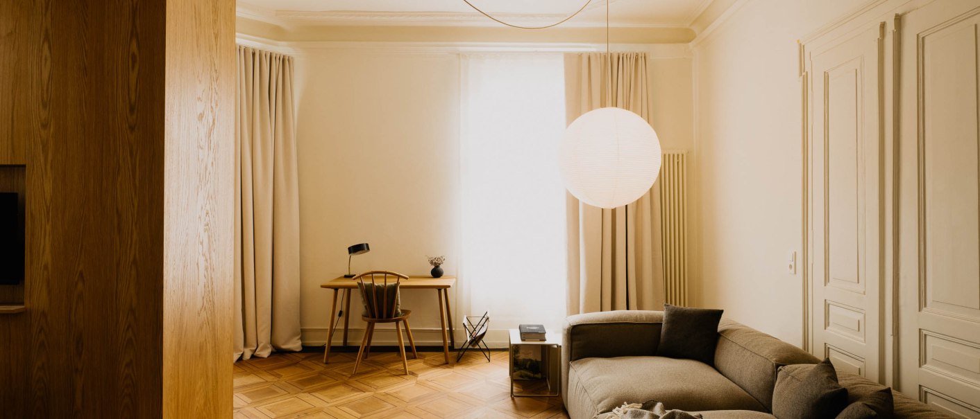Elegantes Wohnzimmer mit einem grauen Sofa, Holzboden, Schreibtisch und gro&szlig;er runder H&auml;ngelampe. Helle Vorh&auml;nge und minimalistische Einrichtung., &copy; Peter Oliver Wolff / DAS EDITH