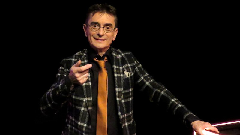 Ein Mann in kariertem Anzug und orangefarbener Krawatte steht auf einer Bühne und zeigt mit dem Finger nach vorne., © Renitenztheater Stuttgart e.V. Ein Mann in kariertem Anzug und orangefarbener Krawatte steht auf einer Bühne und zeigt mit dem Finger nach vorne., © Renitenztheater Stuttgart e.V.