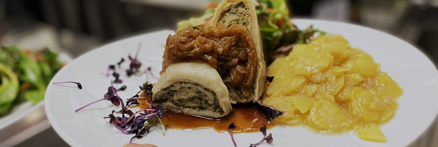 A plate of Swabian Maultaschen, garnished with onions and potato salad, is held in one hand. A kitchen can be seen in the background., &copy; Speisekammer West