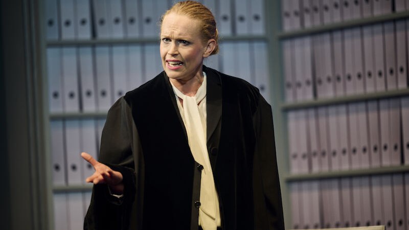 A woman in a black lawyer's coat gestures in front of a shelf full of files. She looks committed and focused., &copy; Theaterhaus Stuttgart e.V.