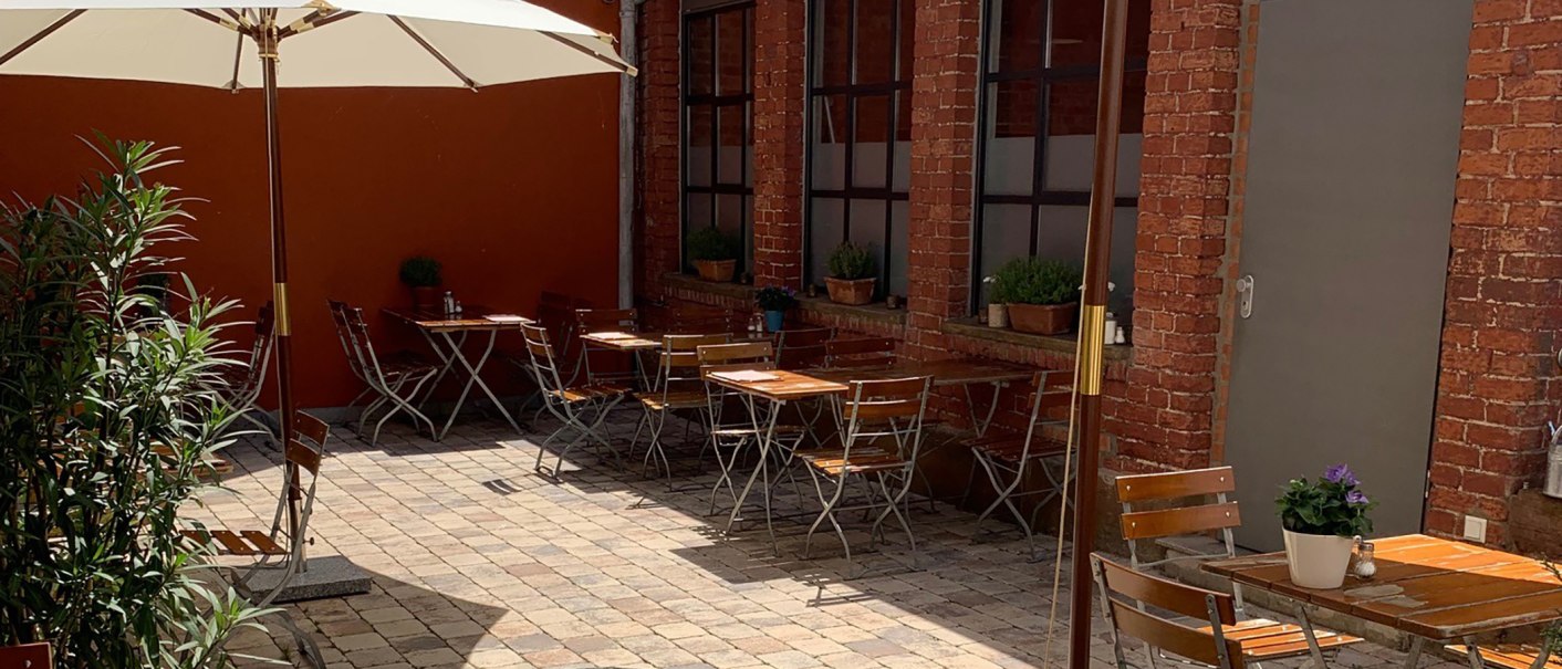 Cozy courtyard with wooden tables and chairs, parasol and plants. Red brick walls and paved floor create an inviting atmosphere., © Gustav Gastro GmbH