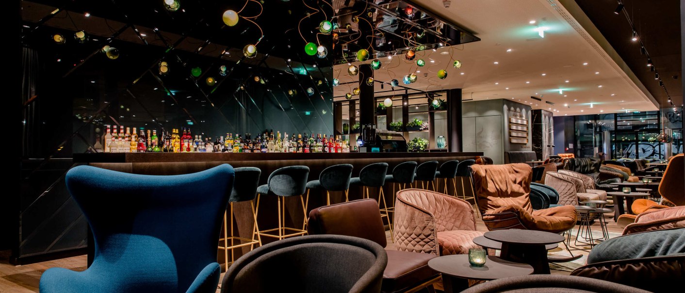 Elegante Bar mit stilvollen Sesseln und Hockern, dekorativen Lampen und einer gut bestückten Theke. Gemütliche Atmosphäre mit modernem Design., © Motel One - Stuttgart Hauptbahnhof