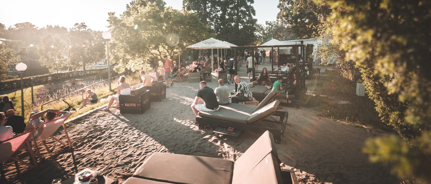 Menschen entspannen an einem sonnigen Stadtstrand in Stuttgart. Liegestühle und Sonnenschirme bieten eine gemütliche Atmosphäre im Sand., © Classic Rock Café Gastronomie- Betriebs GmbH