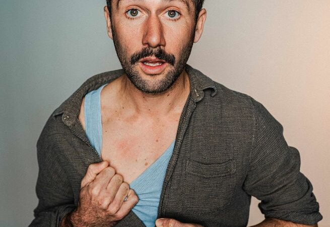 A man with a moustache opens his shirt and points to his heart while holding a microphone. His expression is surprised., &copy; Theaterhaus Stuttgart e.V.