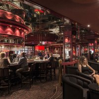 Elegant bar with red décor, mirrors and subdued lighting. Guests sit at the counter and at tables while a bartender serves drinks., © Maritim Hotels Elegant bar with red décor, mirrors and subdued lighting. Guests sit at the counter and at tables while a bartender serves drinks., © Maritim Hotels