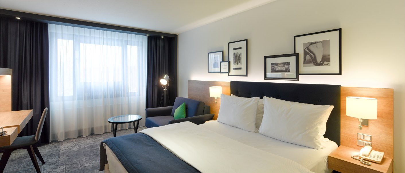 Modern hotel room with double bed, desk, sofa and decorative pictures on the wall. Large windows with curtains let in natural light., © Holiday Inn Stuttgart Modern hotel room with double bed, desk, sofa and decorative pictures on the wall. Large windows with curtains let in natural light., © Holiday Inn Stuttgart